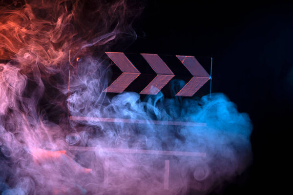 Черно-белая доска для кино крупным планом среди разноцветного красного и синего дыма в руках мужчин дает команду начать съемку на черном изолированном фоне
