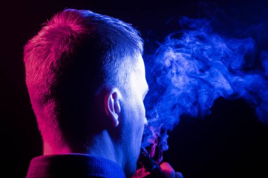 Arkasından bir görünüm bir başının etrafında bir vape sigara ve exhaling gömlekli adamın sağ tarafa bir siyah mavi kendisinden çok renkli duman izole arka plan. İnsan sağlığına zarar