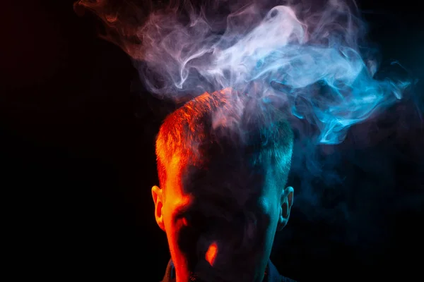 The face of a man in shadow and head is highlighted in red, who smokes a vape and gives out multi-colored smoke of blue and pink above his head, causing harm to his health. The thoughts and soul.