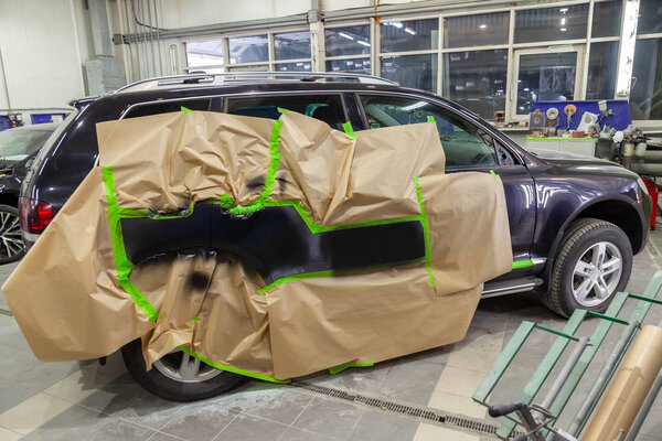 Partial painting of the side elements of the body of a black SUV car after an accident on the road, not damaged parts are covered with paper and tape from getting into paint in a car repair workshop