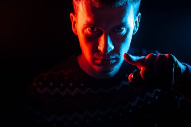 Ciddi bir yüz bir gölge ile kısa saçlı bir adam portresi renkli bir arka ışık mavi ve kırmızı noktaları ile kazak Görüntüleyici, parmağını Sam Amca bir siyah izole arka plan üzerine giymiş.