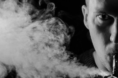 Adam ciddi bir yüz bir gölge ile yarım bir parçası bir vape sigara ve beyaz siyah Yalıtılmış zemin farklı yönlere duman exhaling monokrom renkli bir arka ışık ile portresi