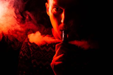 Ciddi bir yüz bir gölge ile bir adam portresi bir vape sigara ve exhaling orenge ve kırmızı renkli bir arka ışık ile çok renkli duman bir siyah farklı yönlere izole arka plan