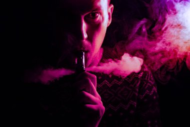 Ciddi bir yüz bir gölge ile bir adam portresi pembe bir vape sigara ve exhaling renkli bir arka ışık ile çok renkli duman bir siyah farklı yönlere izole arka plan