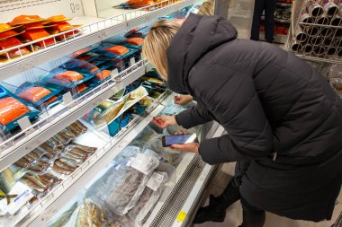 Uzun bir ceket bir genç kız sarışın balık bölümünde alışveriş merkezinde alışveriş yaparken yemek seçer. Sayacın en üst rafta kırmızı taze alabalık.