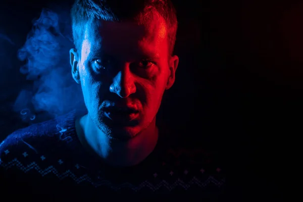 Portrait of a boy from close range who is angry and has bad emotions with a face mash and smoke around on a black isolated background. The face of the guy in the shade with color illumination.
