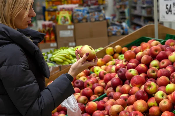一个穿着长夹克的年轻金发女孩在水果部的一家商场购物时 手里拿着一个红绿的苹果 手里拿着很多成熟的产品 — 图库照片