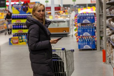Bir genç kız sarışın uzun ceket Gıda Alışveriş arabası ve telefon arasında çok sayıda raf ve sayaçları fırın bölümünde elinde alışveriş merkezinde alışveriş yaparken seçer