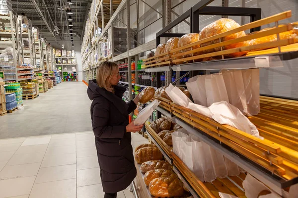 一名身穿长夹克的金发碧眼的年轻女孩在面包店的商场里购物时 在大量的货架和柜台中选择食物 — 图库照片