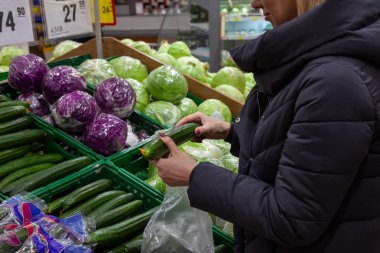 Bir kadın sebze sayaçlarıyla yakınındaki bir süpermarkette bir satın alma işlemi yaptığı: beyaz lahana ve Çin lahanası; ve yeşil bir uzun salatalık seçme elinde tutar ve tazeliğini değerlendirir.