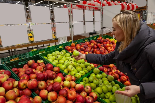 A blond woman in a supermarket buys apples, leans through different types of apples that are beautifully laid out on the shelves to take it, stopping her choice on green, holding it in her hand.