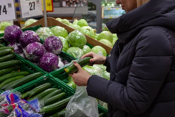 A woman makes a purchase in a supermarket, near the counter with vegetables: white cabbage and Chinese cabbage; and choosing a green long cucumber, holds it in his hands and evaluates its freshness.