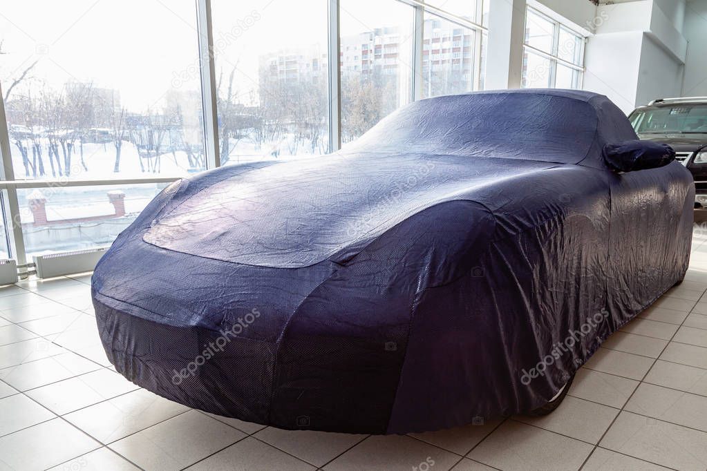 Prepared gift in the showroom, a sports car covered with a blue protective cover awning for cars made of special fabric, sewn in an individual workshop that fits all smooth body lines.