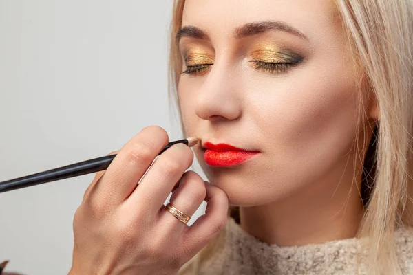 The make-up artist in the studio imposes a luxurious oriental make-up to a young girl blonde, in whose hand she brushes a lipstick of red color, with bright beige-gold and green shades in her eyes.