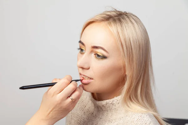 A makeup in a beauty studio, a make-up artist with a brush in her hand puts a product on the lips of a blonde model with a light skin tone and blue eyes, with a contoured cheekbone face profile.
