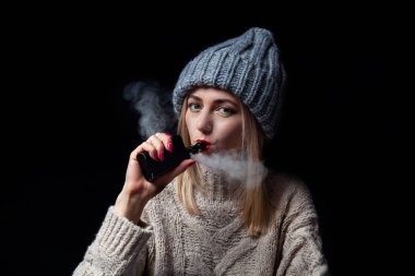 Kız sarışın bir örme kazak ve şapka bir elektronik sigara vape bir manikür ile onun elinde tutan ve dudaklarını kırmızı rujla boyalı ile duman inhales.