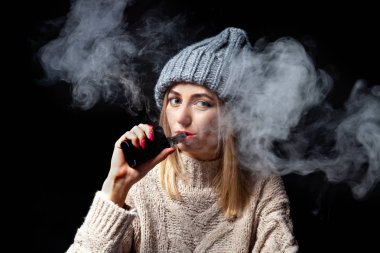 Sarışın bir örme kazak ve şapka bir elektronik sigara vape manikür elini tutar ve onun ağzından duman exhales, siyah bir zemin üzerine kırmızı ruj dudakları boyalı.
