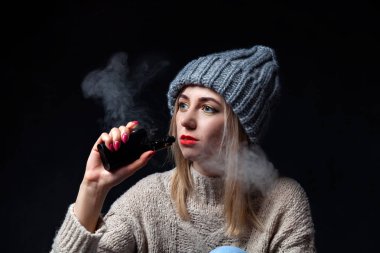 Sarışın bir örme kazak ve şapka bir manikür ile onun elinde bir elektronik sigara vape tutar ve ve mesafe içine görünüyor, dudakları bir arka plan siyah duman kırmızı ruj ile boyalı.