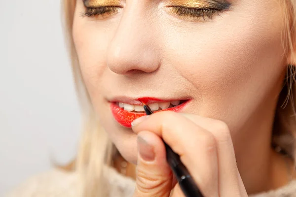 Makeup artist at work in the studio with a brush in his hand imposes a cosmetic product of red lipstick on the lips of the model on which the tone is applied to half.