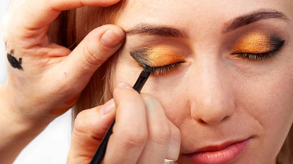 Visagiste modelinin göze makyaj koyarak bir elinde bir fırçayla, öte yandan Doğu tarzı altın ve yeşil gölgeli göz kapağı derisi çeker..