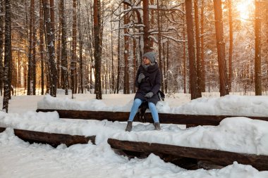 Genç sarışın bir örgü şapka, eşarp ve eldiven arka planı çok ağaçlar ve güneş batımında karla kaplı kış ormandaki bir rüzgârla oluşan kar yığını üzerinde otururken kahve veya çay içer