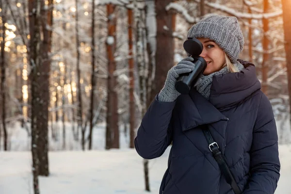 Örme şapka, eşarp ve eldiven içecek kahve veya çay kış karla kaplı ormandaki ağaçlar ve güneş bir sürü zemin karşı günün batımında genç güzel sarışın