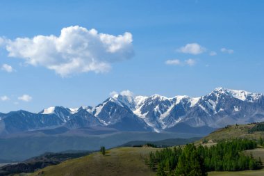 Bir çayır gür yeşil çim ve iğne yapraklı ağaçlar karla kaplı tepeler, mavi bir sis ve beyaz bulutlar dağ taş ridge önünde uzanan ile.