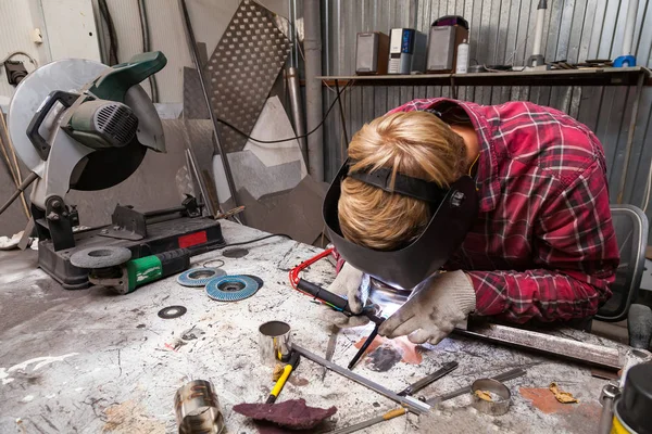 Young guy welder in a checkered red shirt welds a stainless stee