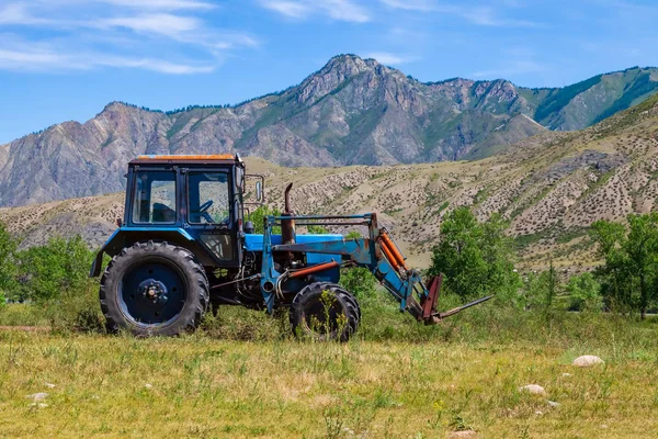 An old blue tractor excavator stands in a field with green and y