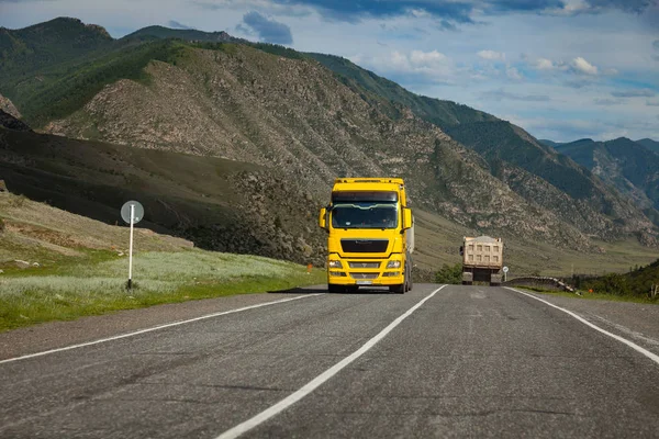 A large yellow truck rides on a highway in the mountains while t