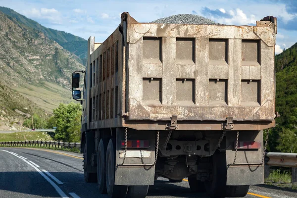 Truck with a gray trailer with rubble in the back of a dangerous highway against the background of rocky mountains and blue sky. Cargo delivery by a transport company.