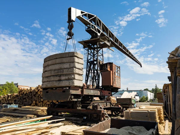 Sawmill with boards and sawn trees near a large old crane with rust under a blue sky with white clouds in summer.