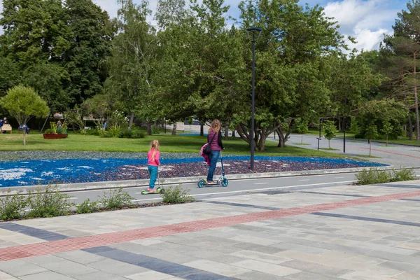 The family spends the weekend in the park. Mom and daughter ride electric scooters on a dedicated bike path near the tiled sidewalk. Modern gadget for fitness and sports.