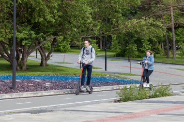 Novosibirsk, Rusya - 06.06.2020: Evli bir çift hafta sonunu parkta geçiriyor, bir erkek ve bir kız, yeşil ağaçlar ve yeni kaldırımlar arasında özel bir bisiklet yolunda elektrikli scooterlara biniyor. Modern aygıt.