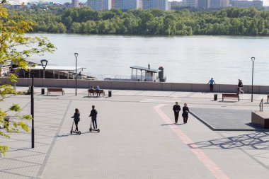 Novosibirsk, Rusya - 06.06.2020: İnsanlar nehrin arka planı, iskele ve yeşil ağaçlar, elektrikli scooterlarla gezen genç bir çift, kaldırım kiremitleri ile kaplanmış kaldırım boyunca yürüyor.