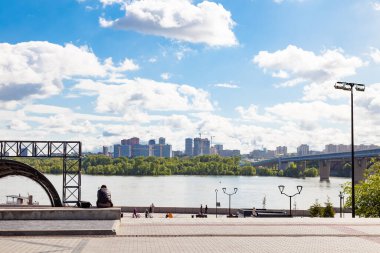 Bir adam bankta oturur ve tepeden tırnağa manzarayı, üzerindeki metro köprüsünü, karşı kıyıda inşaat halindeki evleri, Novosibirsk 'te mavi gökyüzüne ve beyaz bulutlara karşı bir yaz gününü gözlemler..