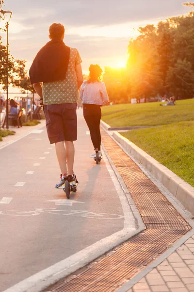 Couple of Teenagers ride on electric scooters on Mikhailovskaya Embankment, many people walk in the park on weekends at sunset on a warm summer day.