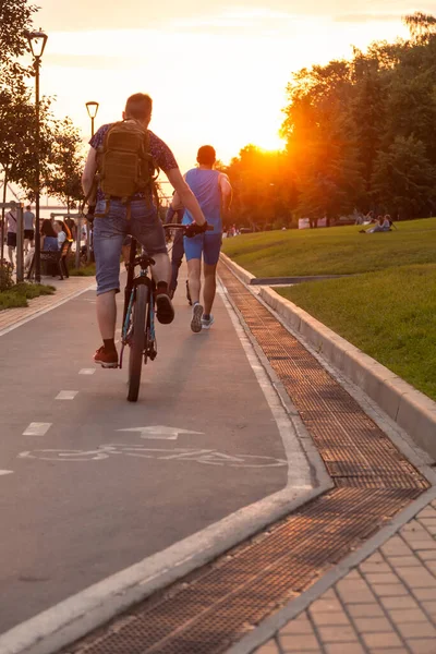 Men run and ride a bicycle along a cycle path on the embankment of Novosibirsk in the park on weekends at sunset on a warm summer day.