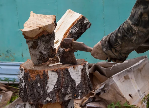 A log split in half with an ax on a deck with male hands in cloth gloves and a camouflage jacket in the backyard. Household work to prepare for the winter.