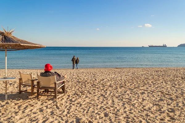 Winter sea beach and calm sea on a sunny day. A lonely  elderly woman sits in a deck chair facing the sea. A loving couple stands by the water. Cargo ship in the distance on the horizon. The concept of the transience of time.