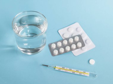 Bir bardak temiz içme suyu, hapları ile iki kabarcık ve mavi bir arka plan üzerinde bir cıva termometre. Hasta olmak için hap almak veya tedaviyi. Sağlık ve ilaç kavramı. Yukarıdan görüntülemek, alan Kopyala.