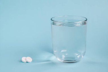 Şeffaf bardak içme suyu ve iki beyaz hap mavi bir arka plan üzerinde. Hasta olmak için hap almak veya tedaviyi. Sağlık ve ilaç kavramı. Ön görünümü ve kopya alanı.