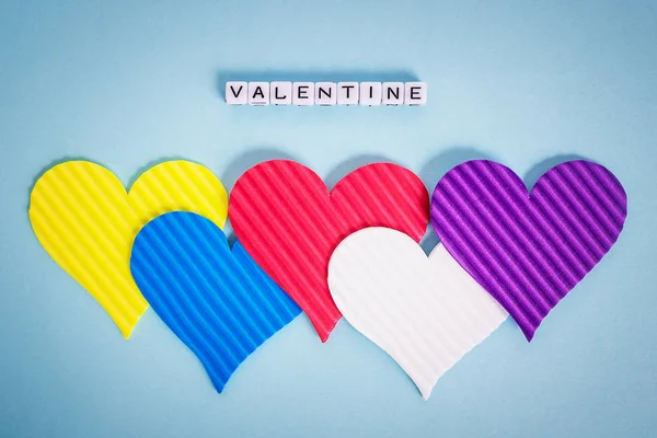 Five multicolored valentine hearts and the inscription Valentine made of white alfphabet cubes on a pastel blue background. Concept of LGBT, love and valentine\'s day. Flat lay, top view.