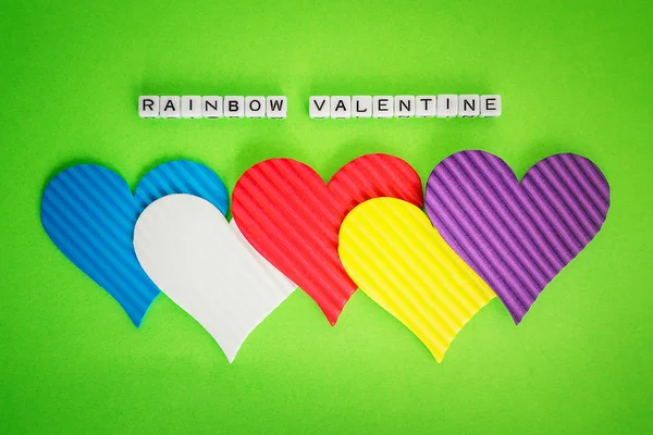 Five multicolored valentine hearts and the inscription Rainbow Valentine made of white alfphabet cubeson a green background. Concept of LGBT, love and valentine\'s day. Flat lay, top view.