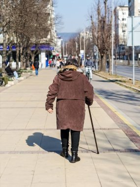 Varna, Bulgaristan, 20 Şubat 2019. Yaşlı bir kadın kahverengi bir ceket giymiş ve bir şehir güneşli bir günde sokak boyunca yürüyüş onun elinde bir sopa ile Arkadan Görünüm.