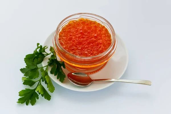 A glass jar of red caviar on a white porcelain plate on a white background. Salmon caviar. Sea fish delicacy. View from above