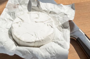 Camembert tüm baş ve pencereden bir güneş ışığı altında bir bıçak açık kağıt paketi. Kahverengi ahşap arka planda yenilebilir beyaz kalıp ile kaplı yumuşak peynir.