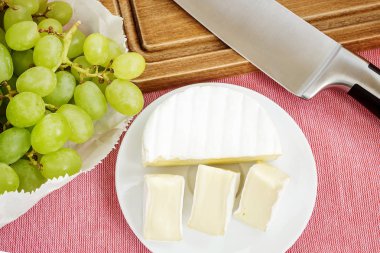 Pembe masa örtüsü üzerinde beyaz plaka ve tatlı yeşil üzüm üzerinde lezzetli peynir Camembert parçaları. Yenilebilir beyaz kalıp ile kaplı yumuşak peynir.