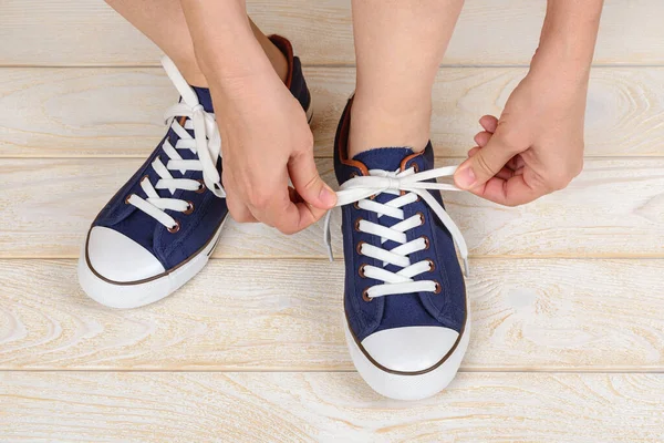 Caucasian woman hands tie white elastic laces on classic blue sneakers or gumshoes indoor. Comfortable shoes for fitness and sport. Shoes with lacing. Top view.