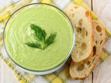 Cam bir kasede taze soğuk salatalık çorbası püresi ve beyaz ahşap bir masada ekmek. Çiğ yemek diyet çorbası Yunan yoğurdu, çiğ salatalık, avokado, dolma biber, dereotu ve sarımsak. Üst görünüm.